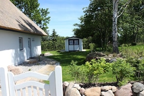 A white building with a thatched roof, a small shed, and a well-maintained lawn with rocks and shrubs. - Holiday home in Usedom with 3 bedrooms (Heringsdorf)