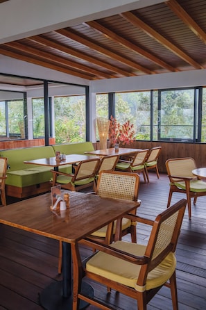 Un restaurante con mesas y sillas de madera, ventanas amplias y vistas al verde exterior.
