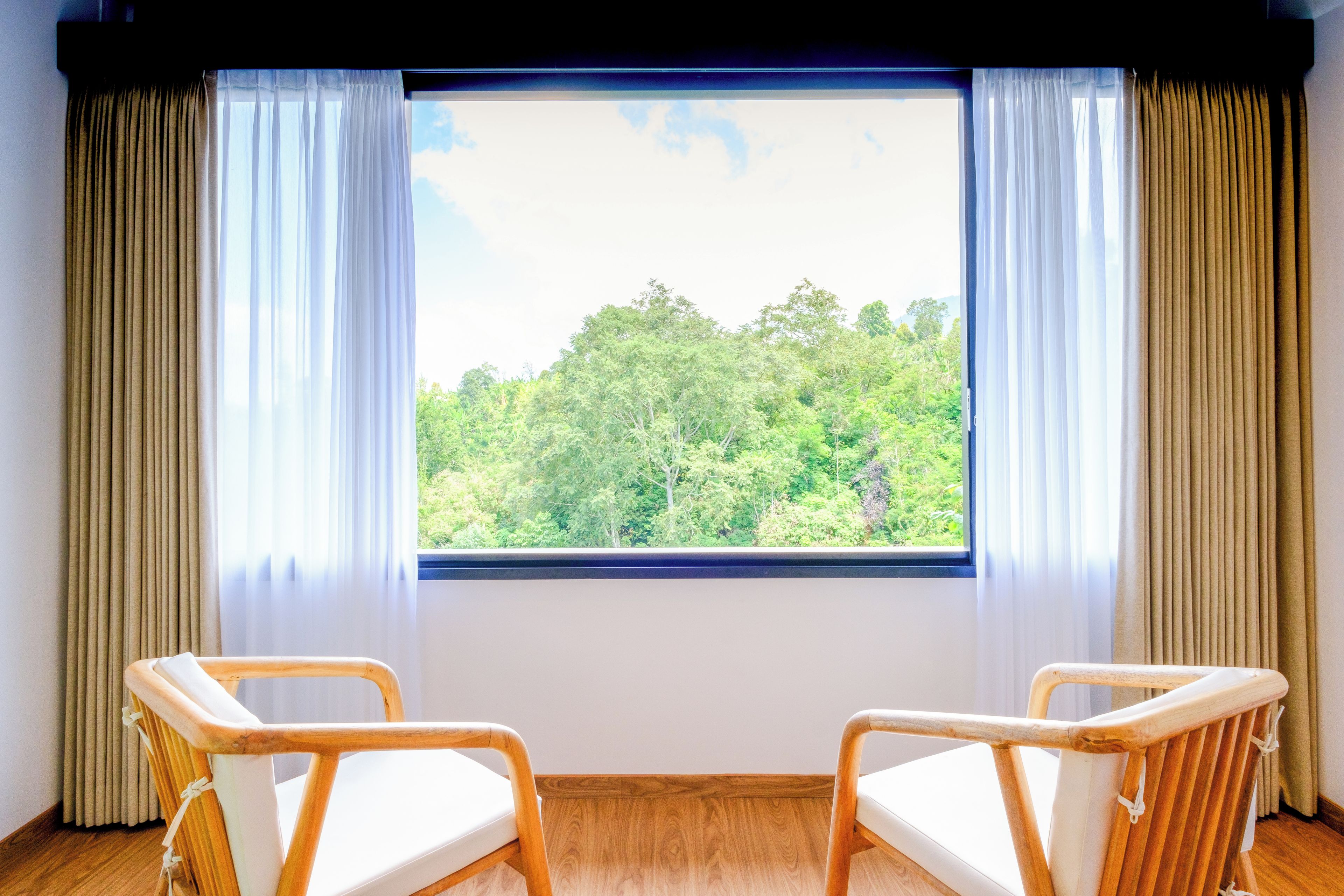 Ein Zimmer mit zwei Holzstühlen, einem großen Fenster mit Vorhängen und Blick auf einen Wald.