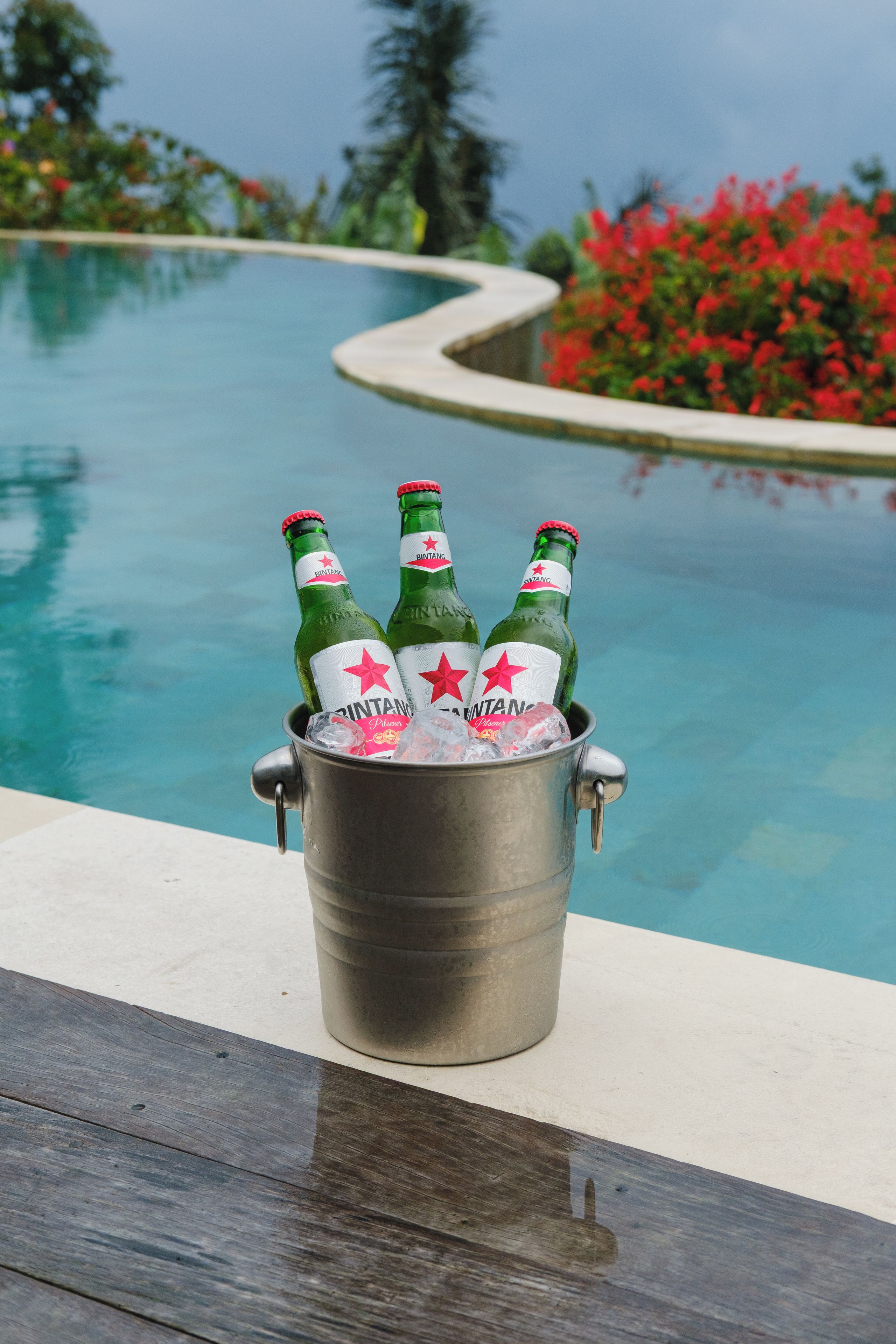 Un balde metálico con tres botellas de cerveza Bintang, colocado al lado de una piscina con vistas despejadas al agua y la vegetación circundante.