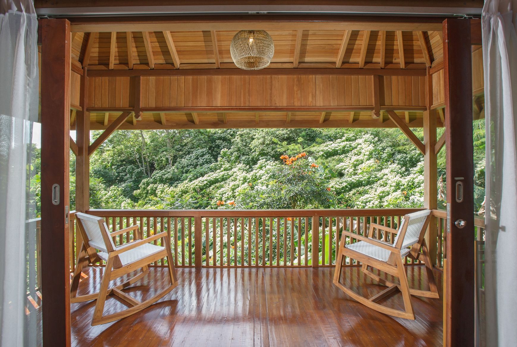 A wooden deck with two rocking chairs, a woven ceiling light, and a view of a lush garden.