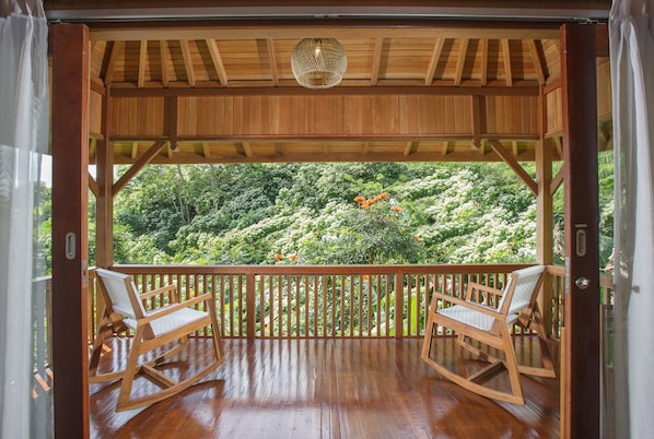A wooden deck with two rocking chairs, a woven ceiling light, and a view of a lush garden.