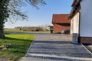A paved driveway leading to a building with a red-tiled roof, a brick exterior, and a grassy area with trees. - Holiday apartment with 2 bedrooms (Bergen \/ Waldhof)