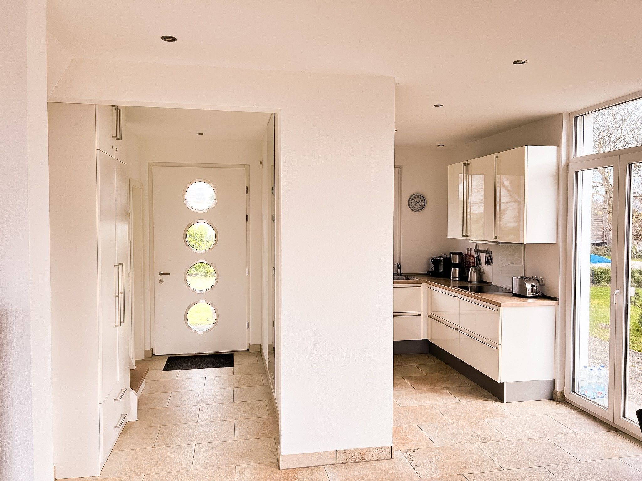 A modern kitchen with a large window, a door with circular windows, and a clock on the wall.