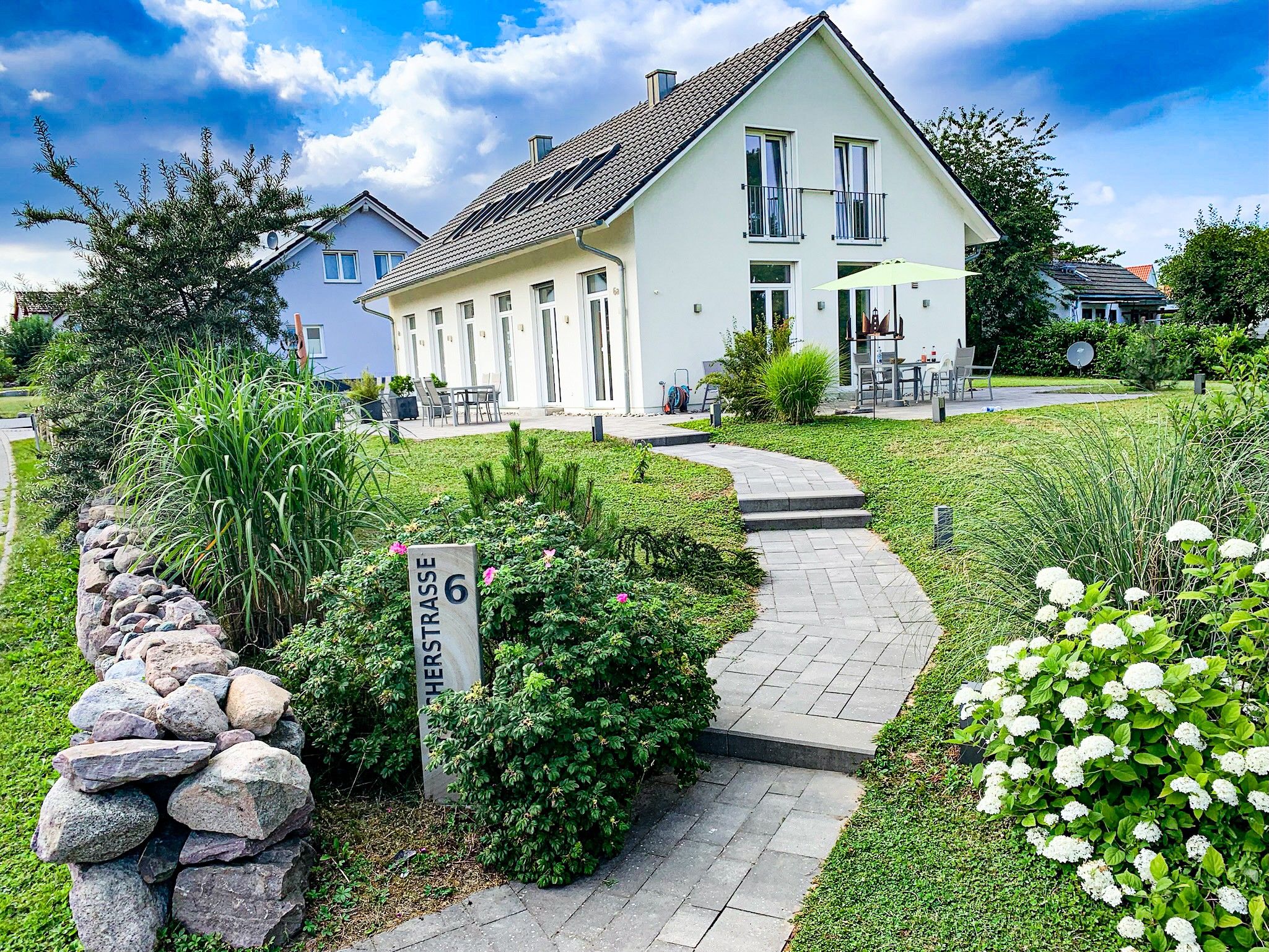 A white house with a paved pathway leading to the entrance, surrounded by greenery and a stone wall.