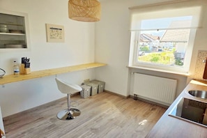 Ett modernt kök med en bänkskiva i trä, en barstol, en hängande lampa och ett fönster med utsikt över ett bostadsområde.