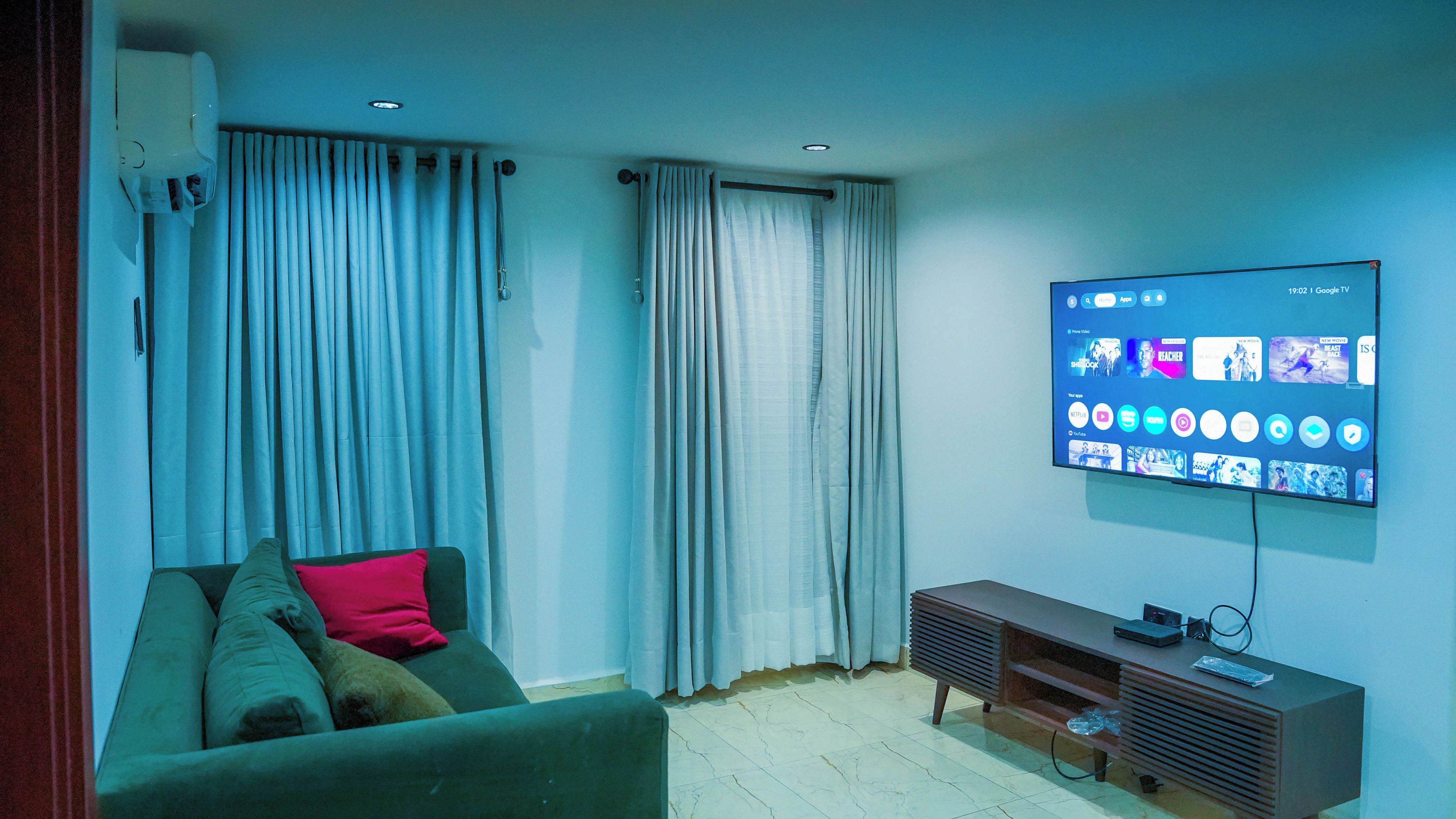 Una sala de estar con una televisión de pantalla plana montada en la pared, un sofá verde con cojines y un mueble de entretenimiento de madera.
