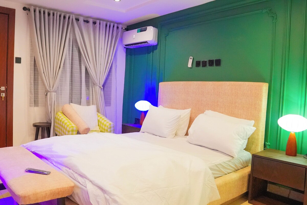 A hotel room with a bed, bedside tables, a nightstand, an air conditioning unit, and a window with curtains.