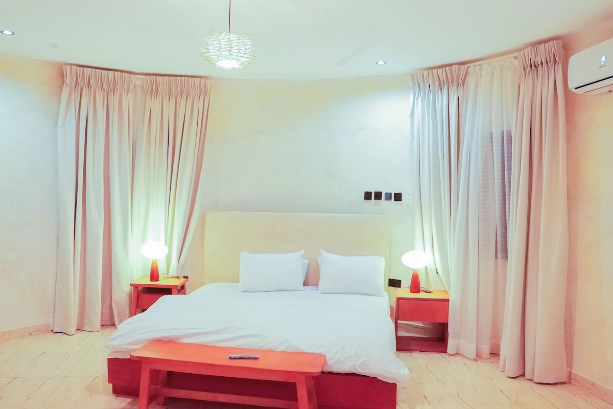 A hotel room with a bed, bedside lamps, an air conditioning unit, and curtains.
