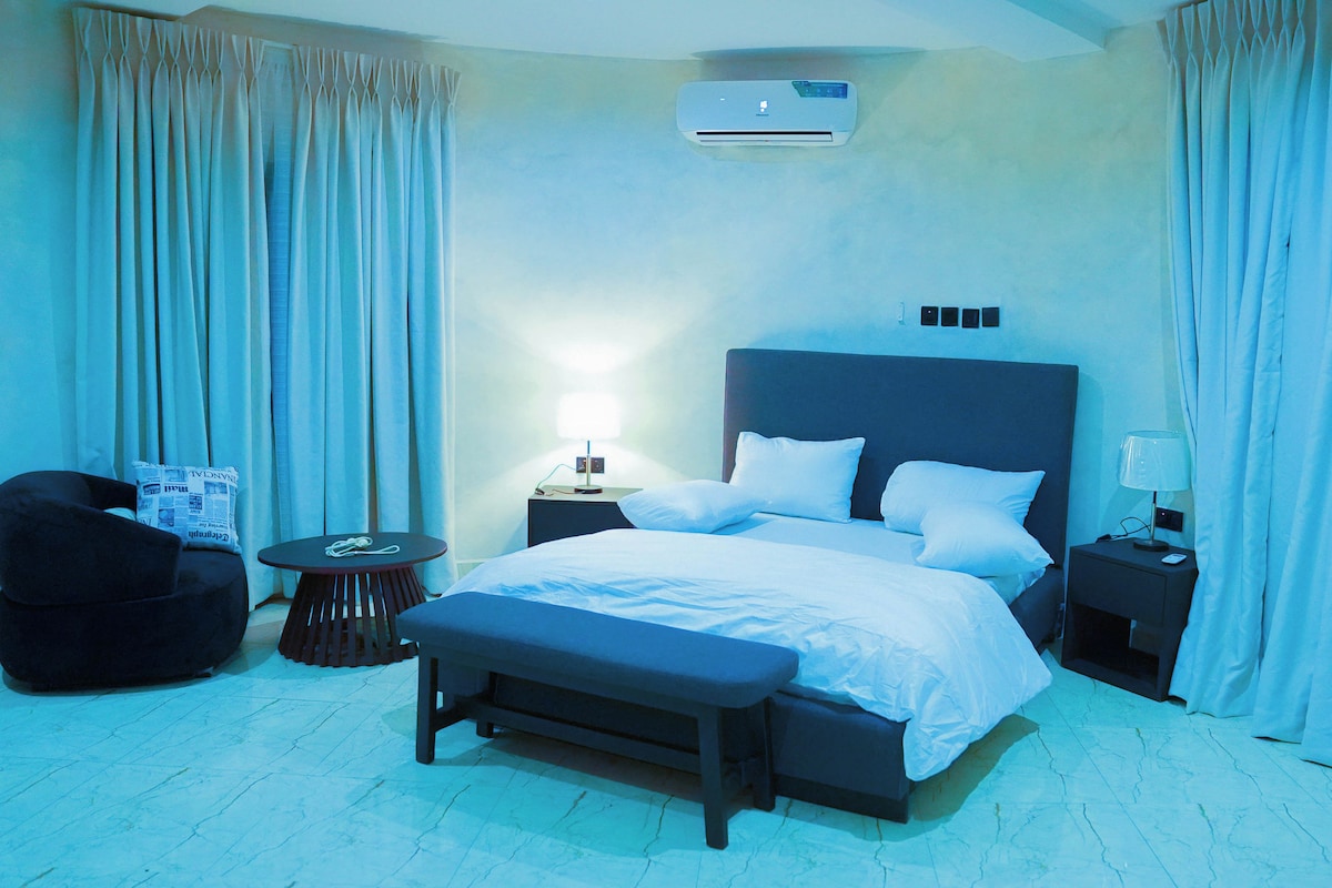 A hotel room with a bed, bedside tables, a bench, a chair, and an air conditioning unit.
