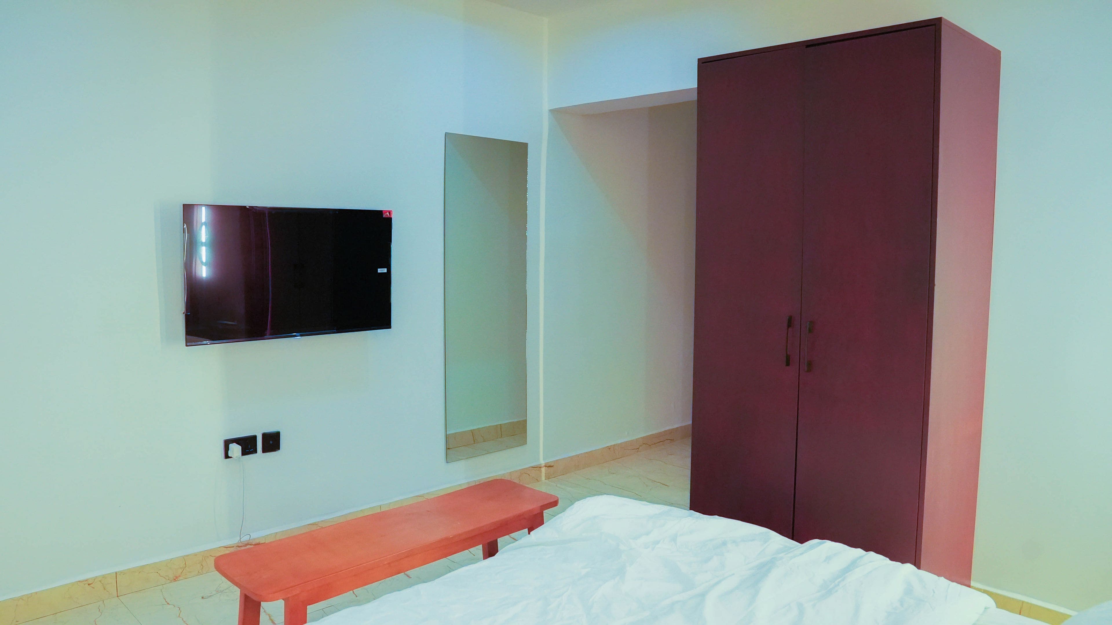 Una habitación con una cama, un banco, un armario y un televisor instalado en la pared.