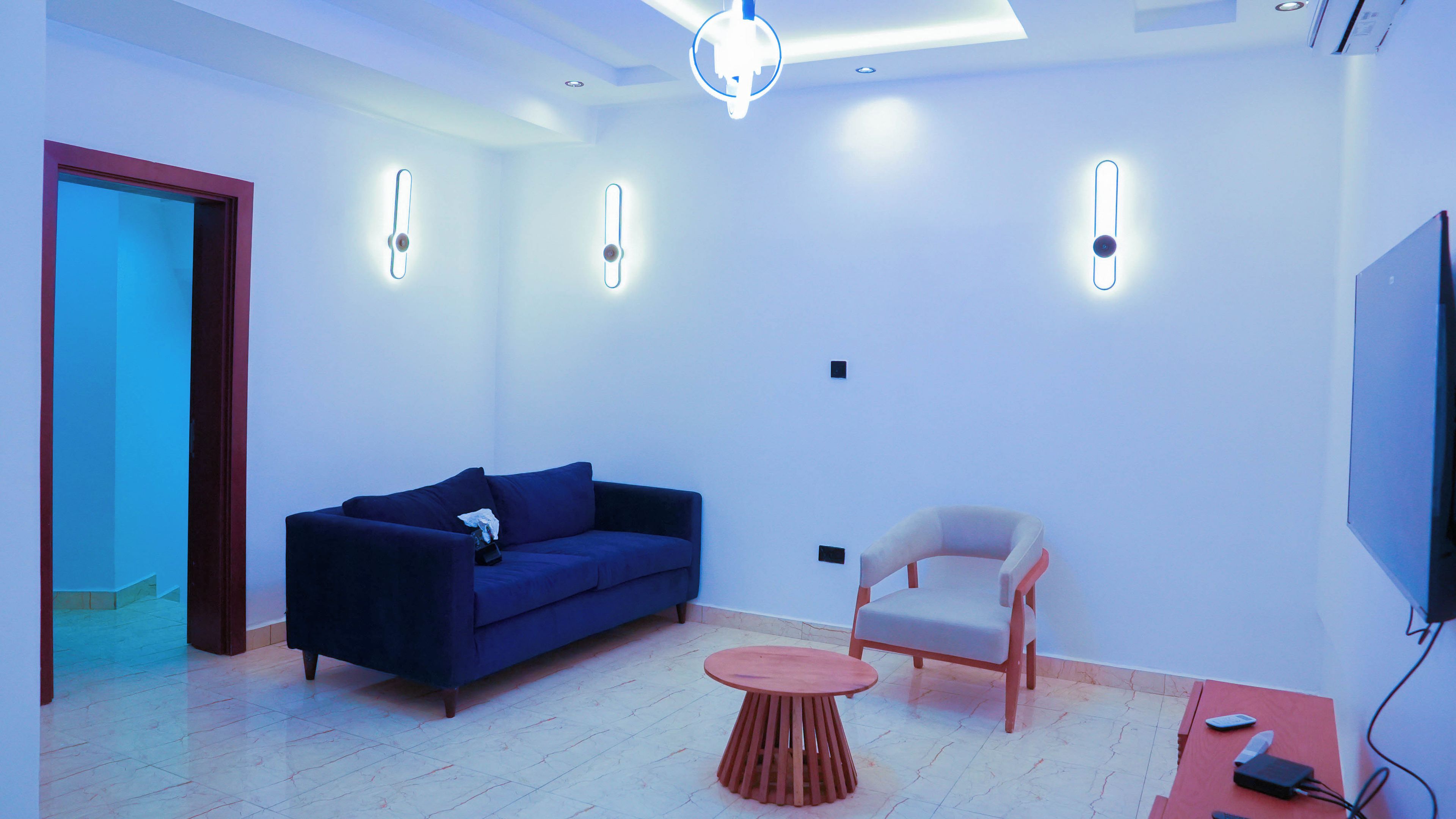 Una sala moderna con un sofá azul oscuro, una silla de madera, una mesita redonda y un televisor de pantalla plana montado en la pared.