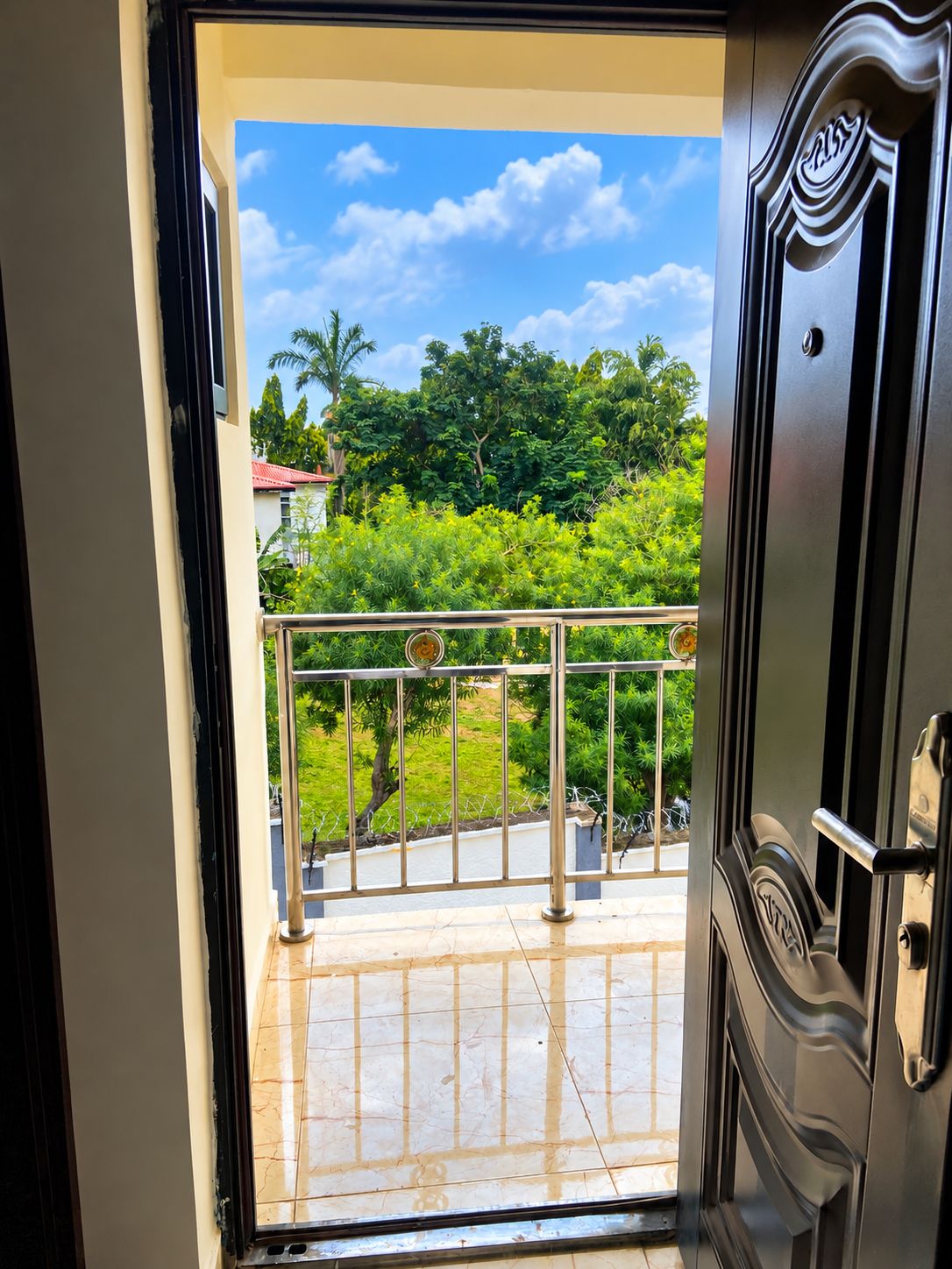 An open door leading to a balcony with a metal railing, a view of greenery, and a clear sky.