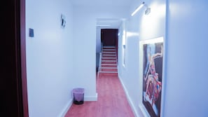 Um corredor com escada que leva a um quarto escuro, uma lixeira e um quadro na parede.