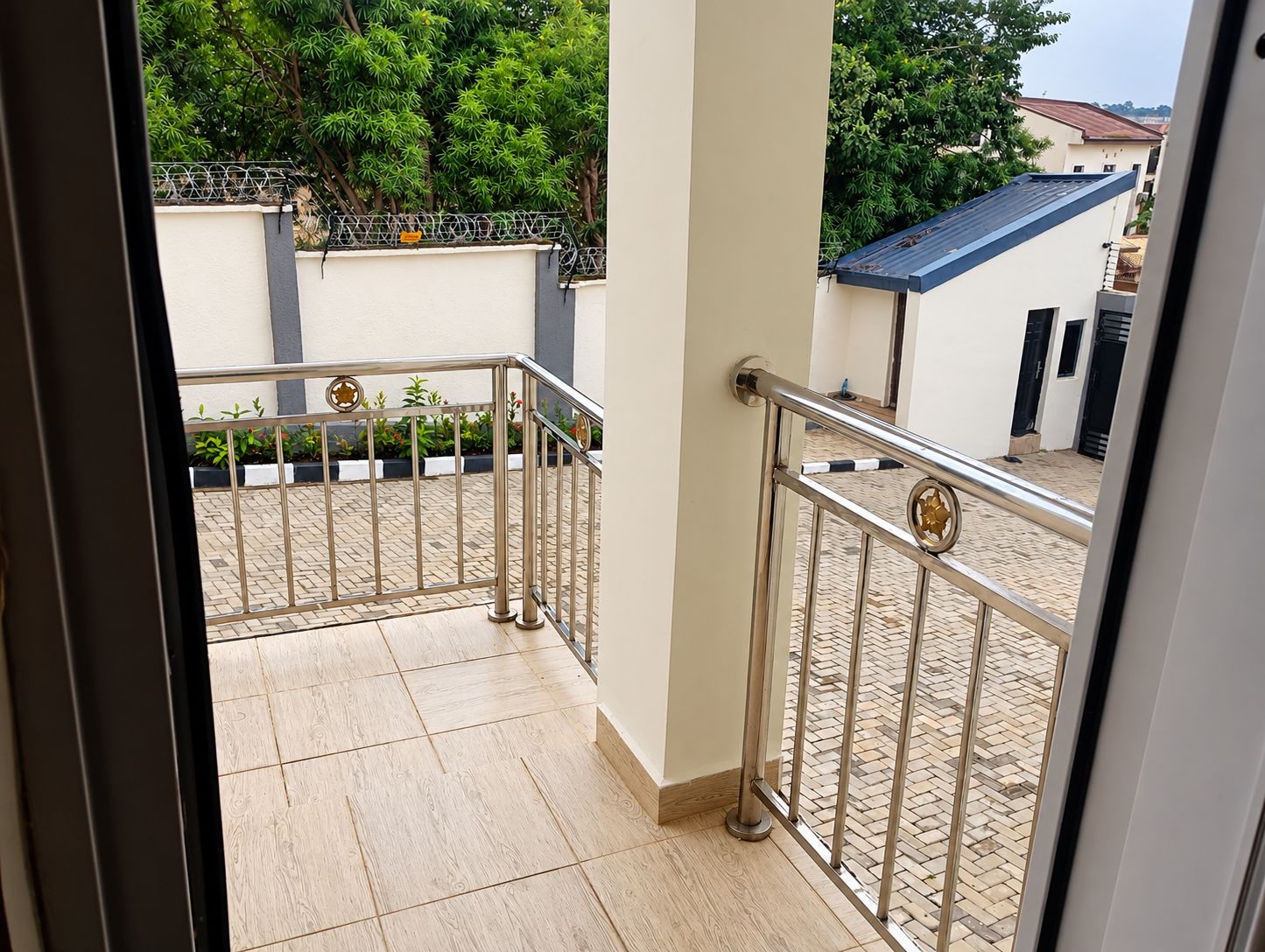 A balcony with a metal railing, a tiled floor, and a view of greenery and buildings in the background.