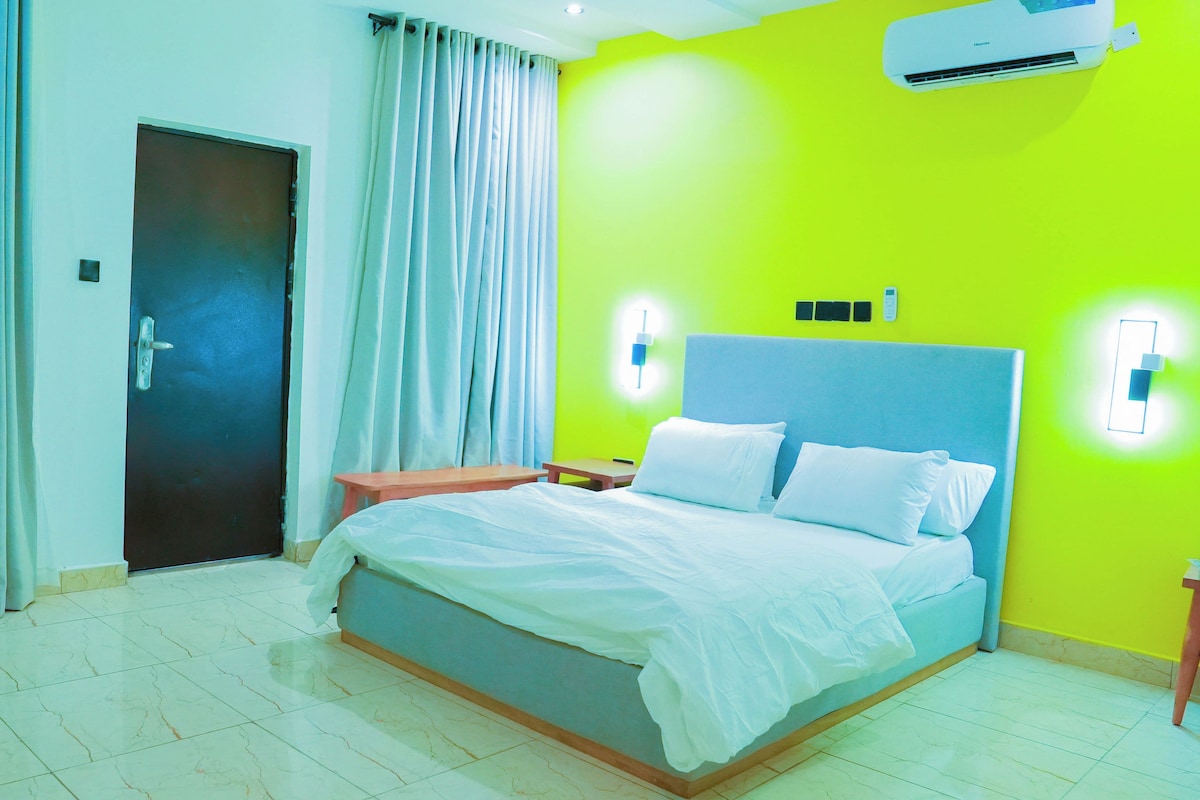 A hotel room with a bed, curtains, a door, a bedside table, a nightstand, and an air conditioning unit.