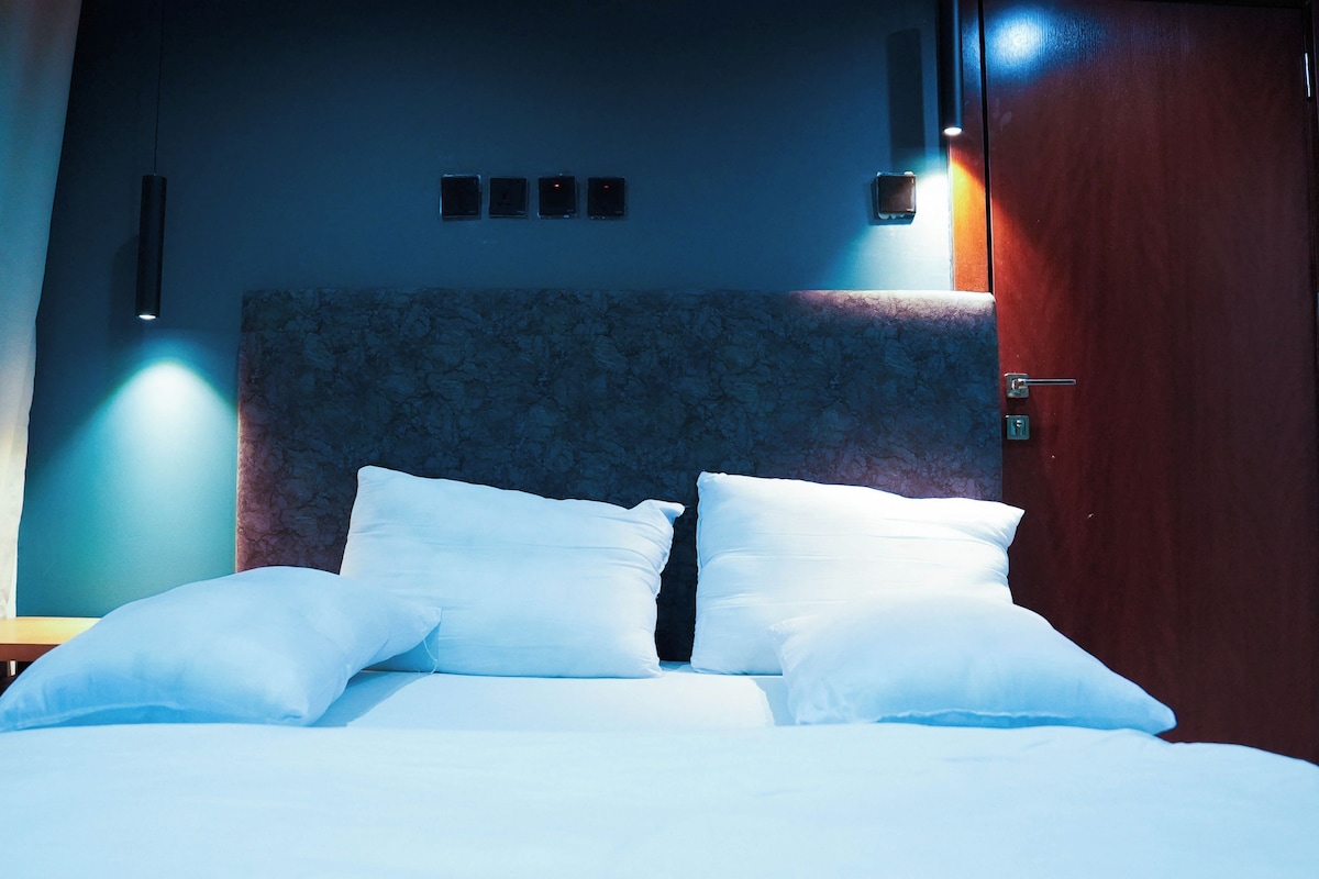 A hotel room with a bed, a nightstand, a wall-mounted light, and a door.
