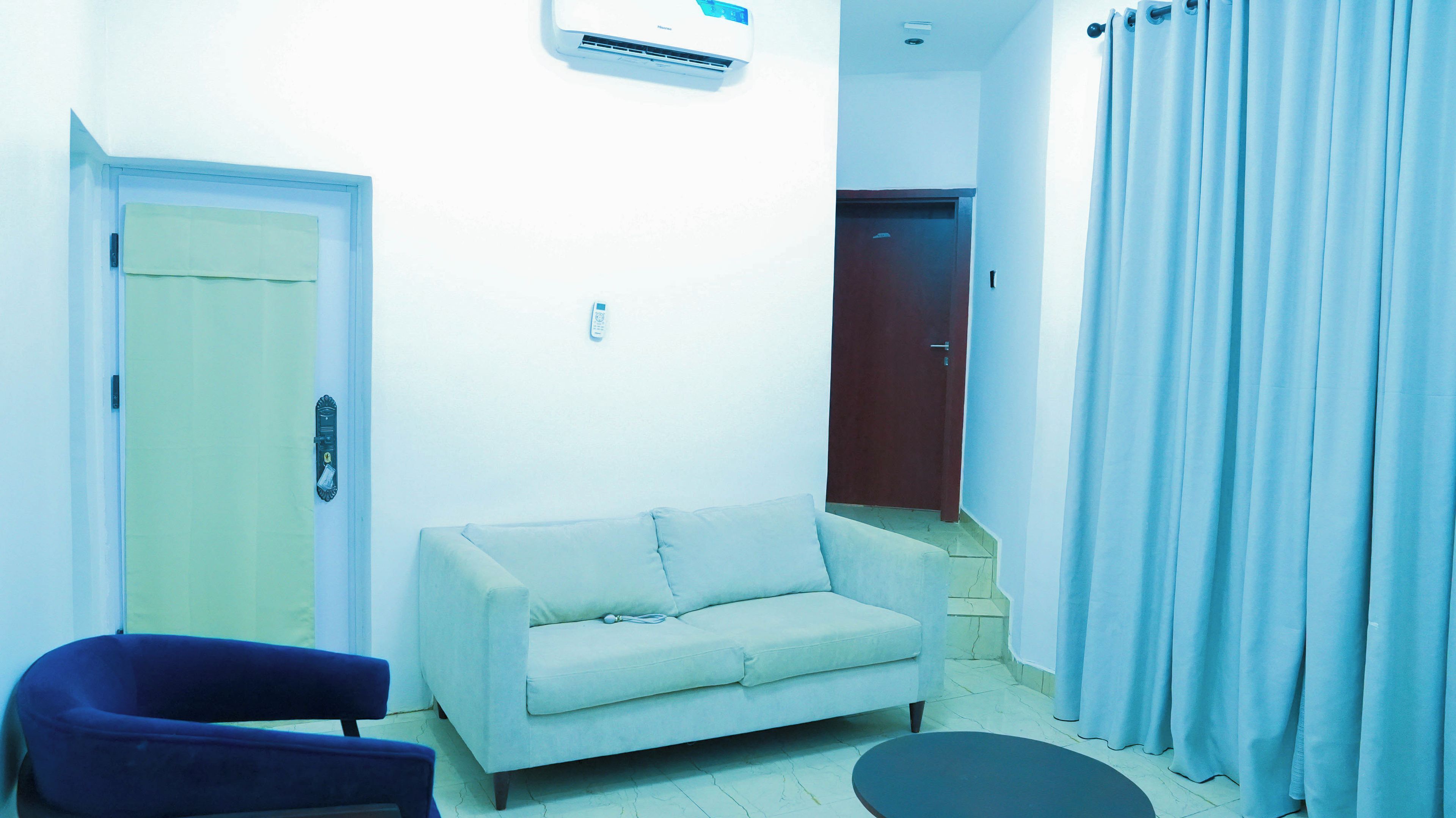A room with a sofa, a chair, a door, an air conditioning unit, and a curtain.