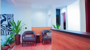 A reception area with a wooden reception desk, two grey armchairs, a small round table, and potted plants. - Bluenix Haven Luxury Apartments  (Abuja Municipal)