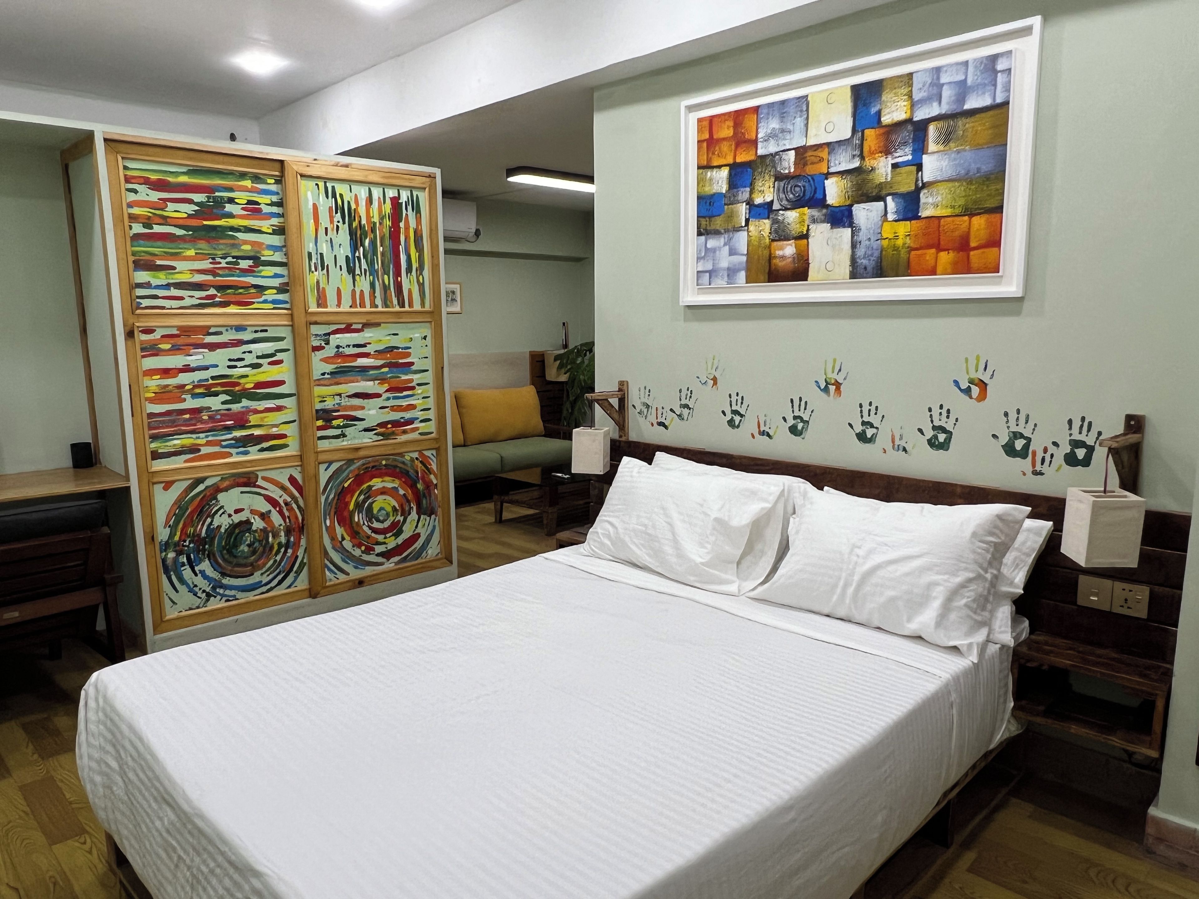 A hotel room with a bed, a wooden partition with artwork, a wall with handprints, and a painting.