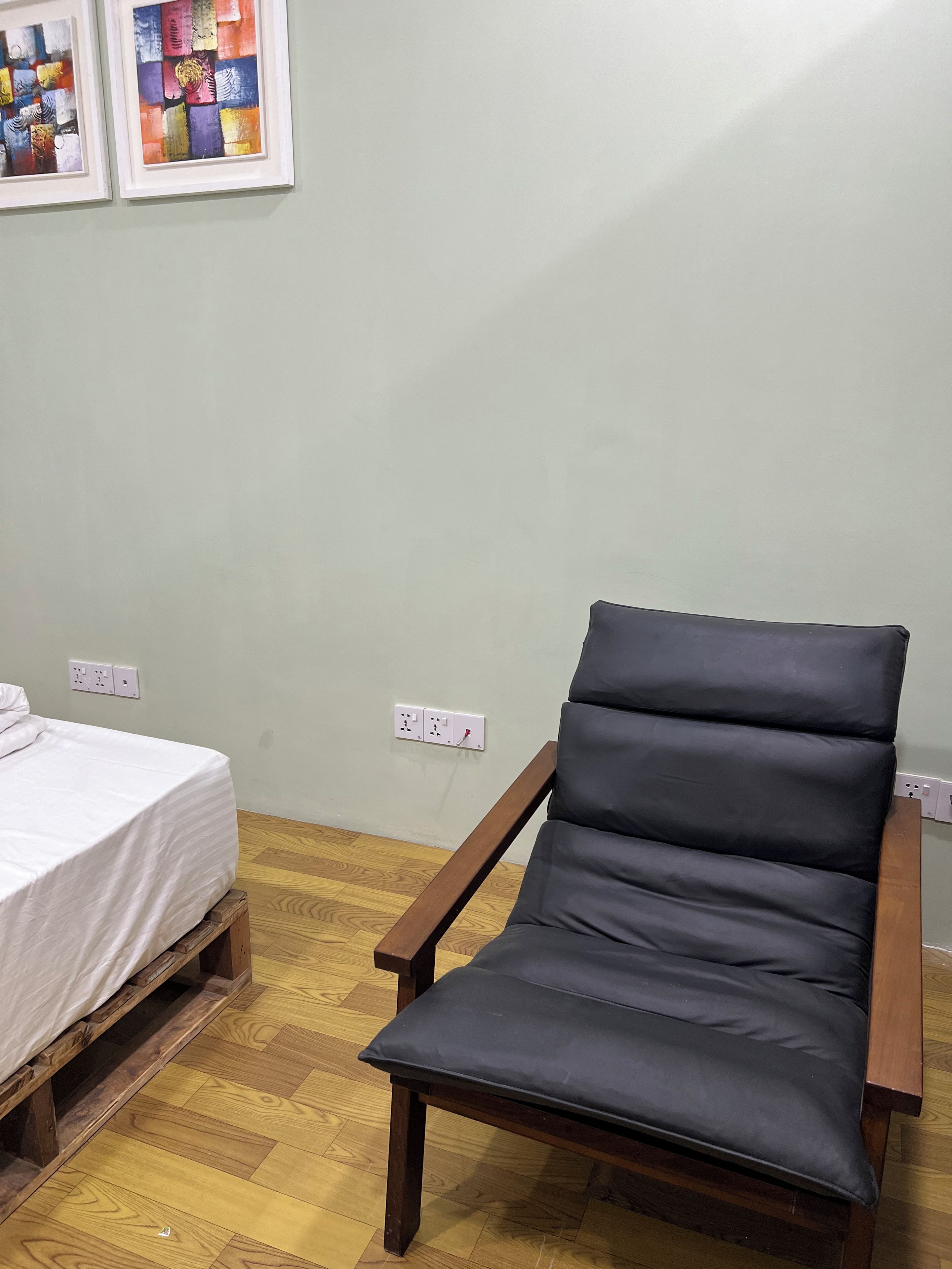 Een kamer met een bed, een houten bankje, een zwarte fauteuil met kussens, en twee kunstwerken in lijsten aan de muur.