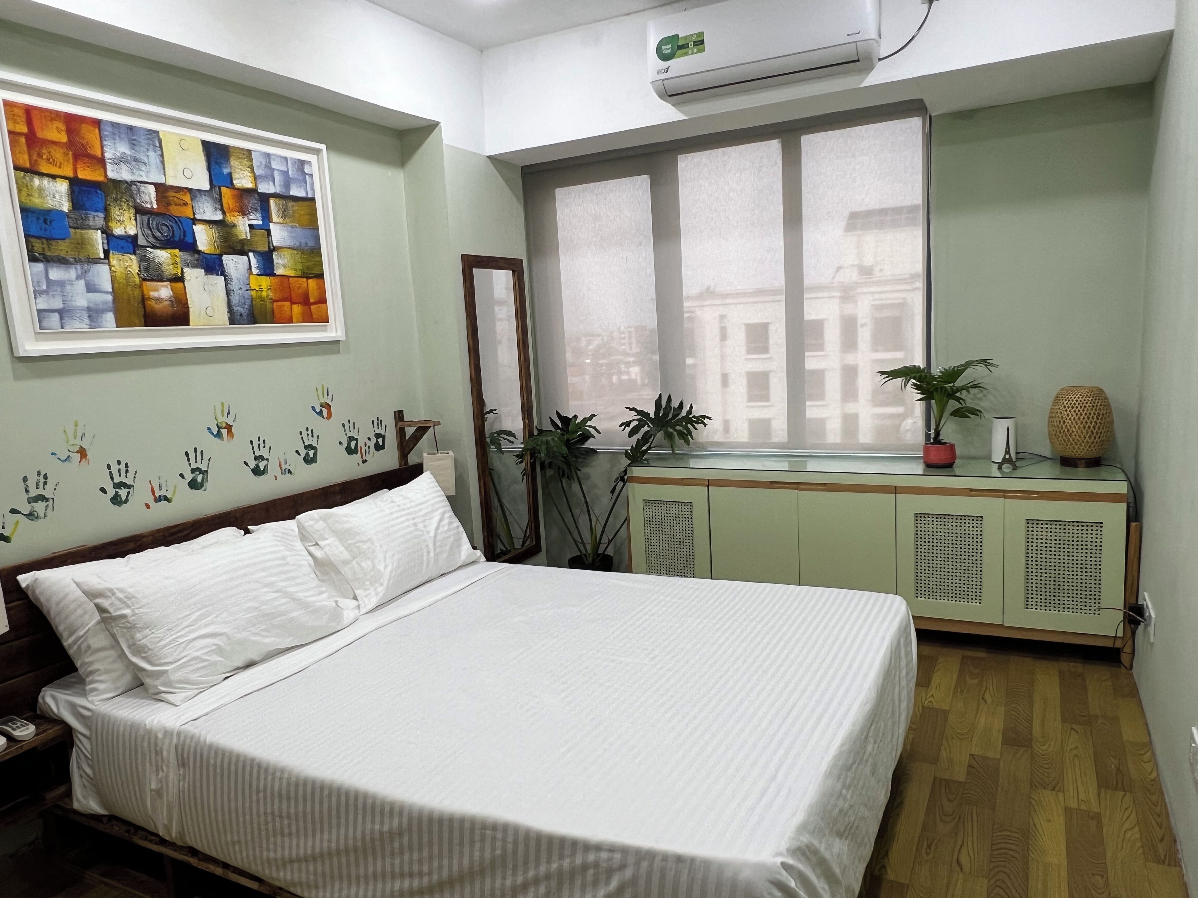 Habitación de hotel con cama, mesita de noche, espejo, ventana con vistas y una pared decorada con una obra de arte colorida.