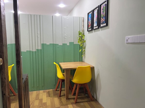 Ein Zimmer mit einem Holztisch, gelben Stühlen, einer grün-weißen Trennwand und gerahmten Kunstwerken an der Wand.