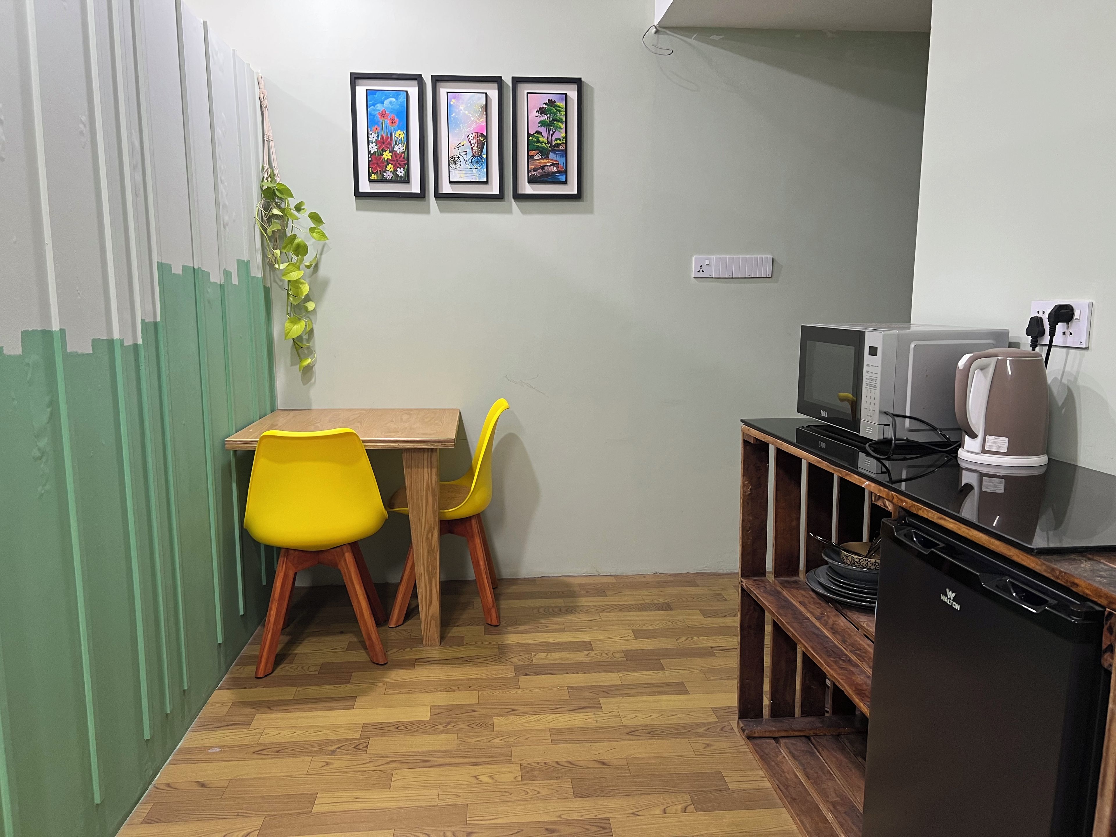 A small dining area with a wooden table and two yellow chairs, a refrigerator, a microwave, and a kettle on a countertop.