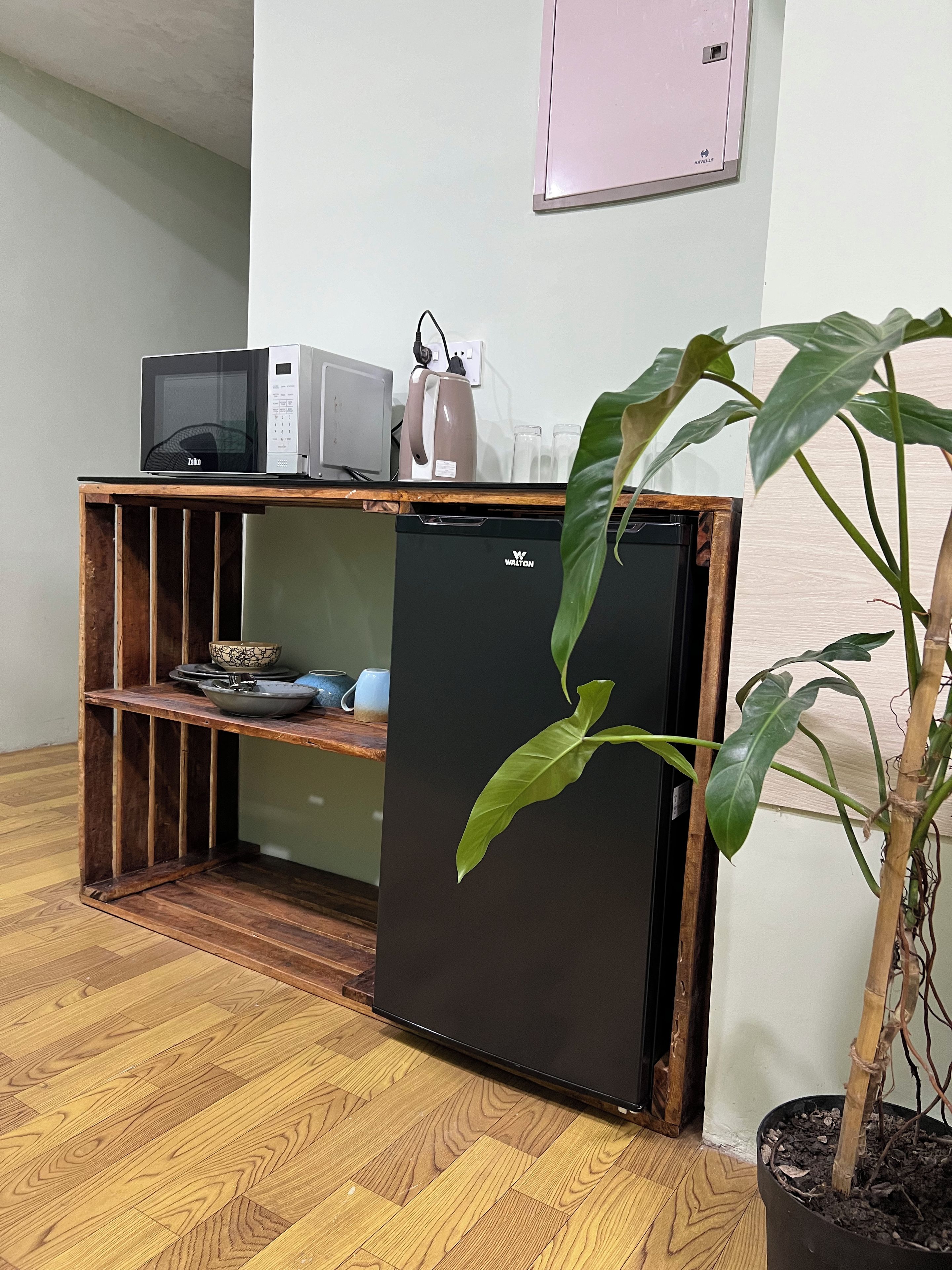 Una kitchenette compacta con microondas, hervidor y un refrigerador pequeño. Una planta en maceta está colocada junto al refrigerador.