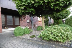 A brick building with a paved entrance, a small garden with shrubs, and a tree providing shade. - Stork's nest Petershagen (Petershagen)