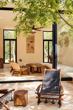 A room with a large window, a hanging plant, a woven chair, a wooden table, and a wall with a hanging artwork. - Casa Xulkan: Stunning Jungle Villa with 2 Private Pools (Tulum)