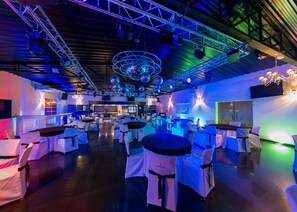 Un espace intérieur vaste et accueillant, doté de plusieurs tables et chaises rondes, d’un bar et d’une scène équipée d’un système d’éclairage.