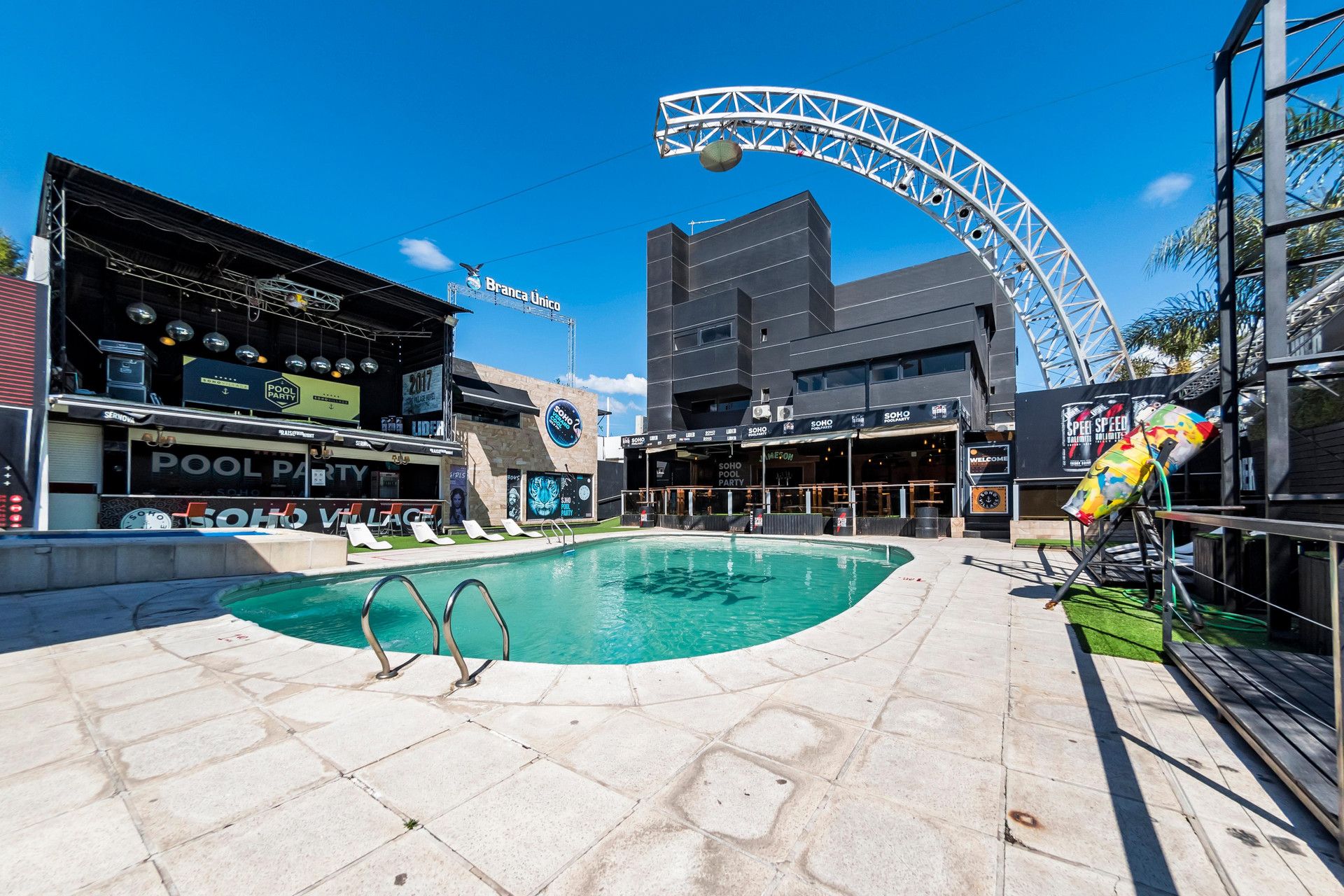 Un espace piscine avec un bâtiment moderne en arrière-plan, une grande structure en forme d’arche et un panneau indiquant qu’une fête est organisée au bord de la piscine.