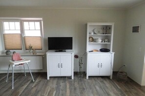 A room with a television on a white cabinet, a white shelf with decorative items, a radiator, and a window with blinds. - Huus Moorstrand holiday apartment "Moor" (Nordstrand)