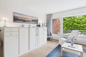 A modern room with a white cabinet, a painting, a window with a view of greenery, and a white chair. - Holiday apartment Dorfschatz (Langeoog)