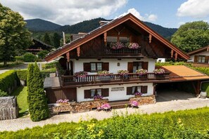 Ein zweigeschossiges Chalet mit Balkon, umgeben von üppigem Grün und Bergen im Hintergrund.