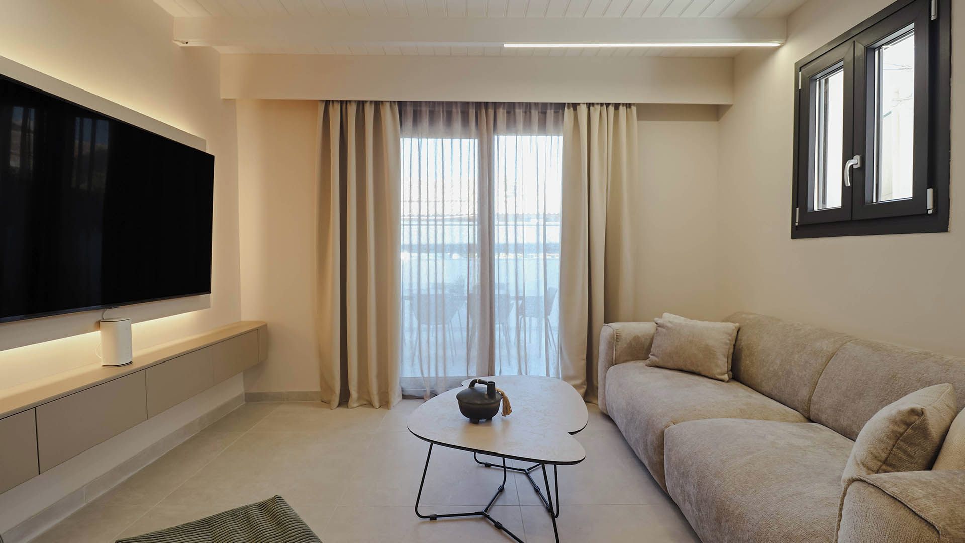 Una sala de estar moderna con un televisor de pantalla plana, un sofá, una mesa de centro y vistas amplias a través de ventanas grandes.