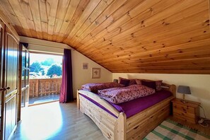 Techo de madera, una cama con colcha morada, una mesita de noche con lámpara y vistas a una terraza y árboles.