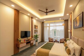Habitación de hotel con cama, televisión, escritorio con computadora, una silla, planta y ventana con cortinas.