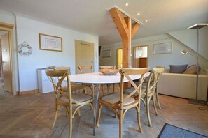 Ein Essbereich mit einem runden Tisch, sechs Stühlen, Holzbalken darüber und einer Couch im Hintergrund.