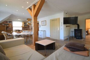 Un salon confortable avec un canapé, une petite table et un téléviseur. Au fond, on aperçoit une cuisine avec des meubles en bois et une table à manger.