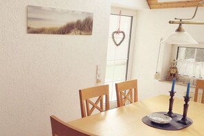 A dining area with a table, chairs, a wall-mounted painting, a heart-shaped decoration, a window with curtains, and a wall-mounted lamp. - KrosseKrabbeNorddeich (Norden-Norddeich)
