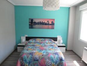 A bedroom with a bed, a cityscape painting, a white bedside table, and a white lamp.