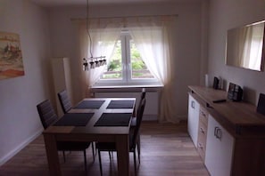 A dining area with a table and chairs, a window with curtains, a radiator, and a wooden cabinet with drawers. - Holiday home with 2 bedrooms (Wurster Nordseeküste)