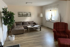 A living room with a sofa, a coffee table, a red armchair, a potted plant, a lamp, and a window with curtains. - Holiday home with 2 bedrooms (Wurster Nordseeküste)
