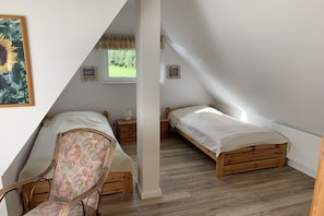 A bedroom with a sloped ceiling, two single beds, a wooden chair, a bedside table, and a window with a view of greenery. - Holiday home with 2 bedrooms (Wurster Nordseeküste)