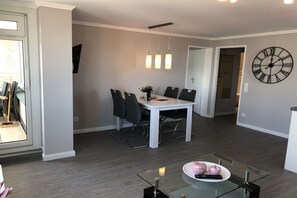 A modern dining area with a white table and chairs, a large wall clock, and a glass table with a bowl of fruit. - Holiday apartment with 2 bedrooms (Westerland)