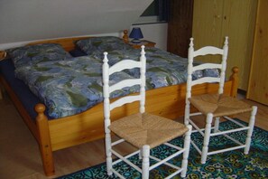 Ein Bett mit gemustertem Überwurf, zwei weiße Stühle mit gewebten Sitzflächen, ein kleiner Tisch mit einer Lampe und ein Teppich mit Muster auf Holzdielen.