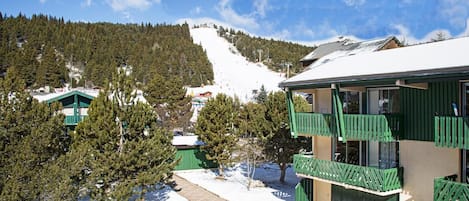 Ein mit Schnee bedeckter Berg mit einer Skipiste, ein Gebäude mit grünen Balkonen und ein klarer, blauer Himmel.