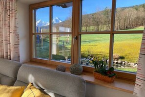 A room with a large window overlooking a green field and mountains. There is a sofa with cushions and a basket of flowers on the windowsill. - Holiday apartment with 1 bedroom (Bischofswiesen)
